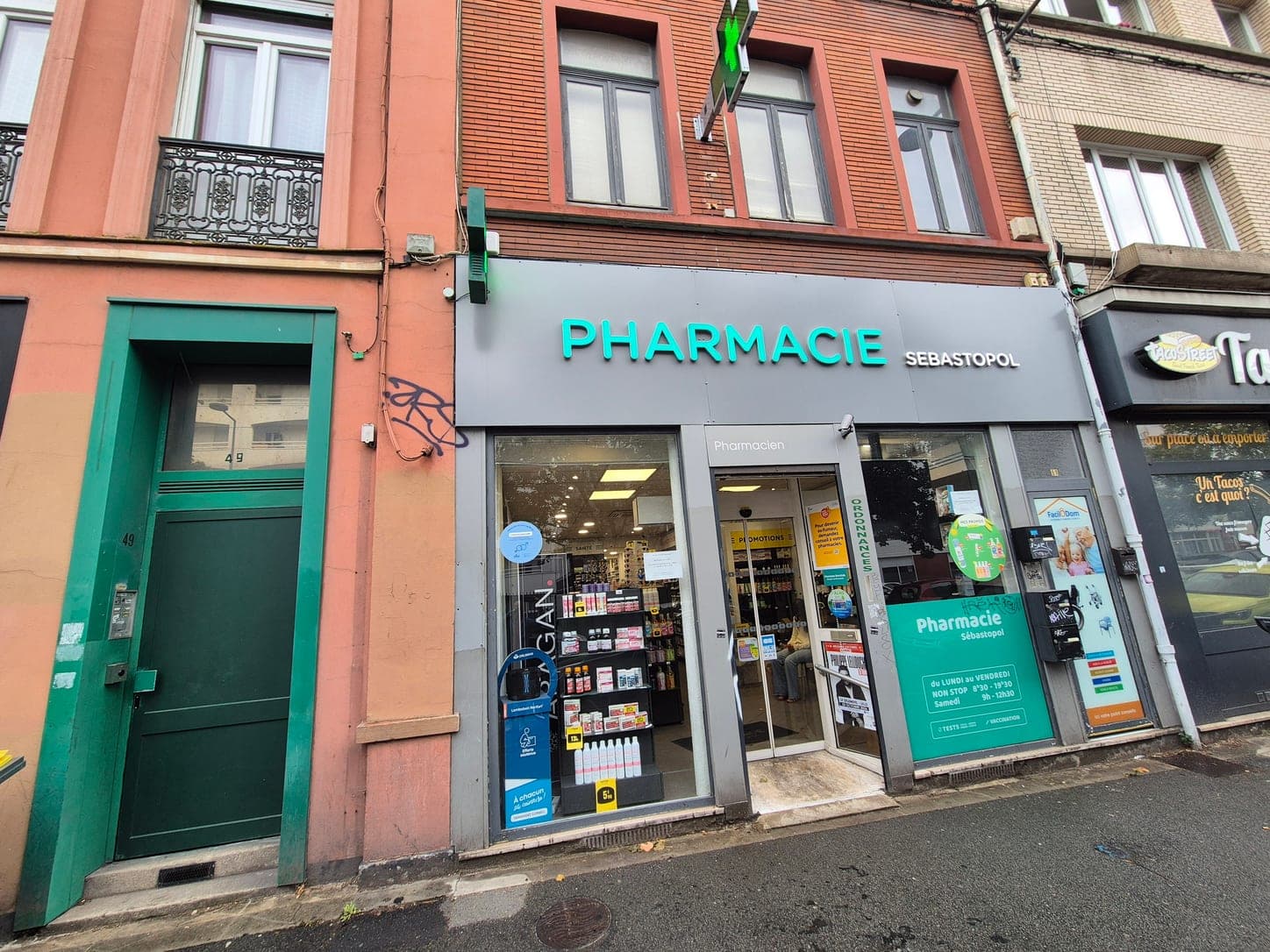 La devanture de la Pharmacie Sébastopol vue en remontant la rue des Postes à Lille, entre une porte verte et les commerces voisins, la croix de pharmacie au-dessus du linteau.